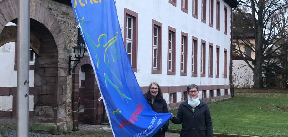 Das Foto zeigt die Bürgermeisterin der Stadt Moringen und die Gleichstellungsbeauftragte vor dem Rathaus unter der Fahne mit der Aufschrift Nein zu Gewalt an Frauen 